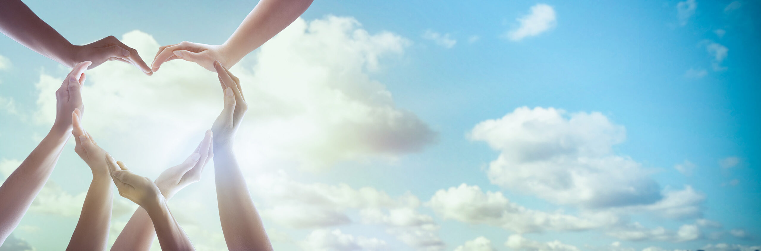 Many hands coming together to form a heart shape, on a sunny background with blue sky and clouds with a silver lining